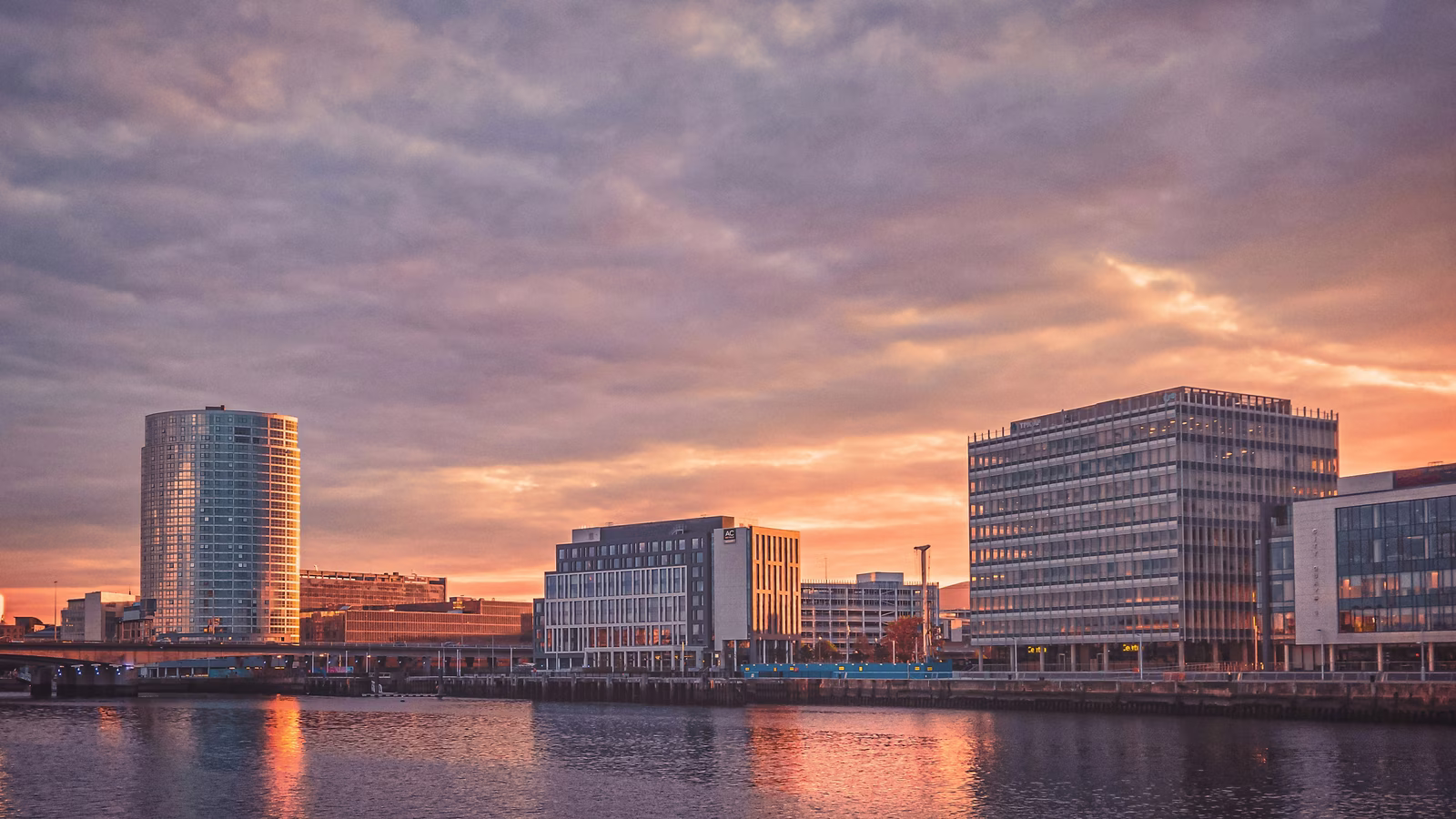 A city skyline at sunset with modern buildings reflected in the water of a river or bay
