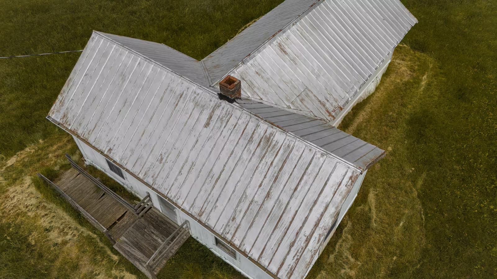 Aerial view of a house with a corrugated metal roof and a chimney, surrounded by green grass.